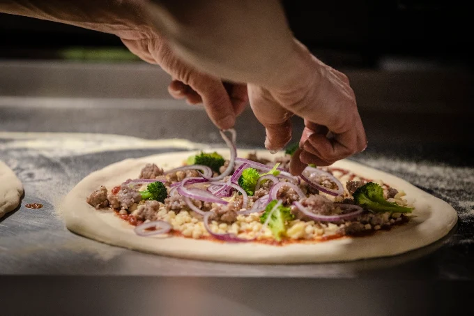 Pizzero de Porco Dio montando a mano una pizza con cebolla roja