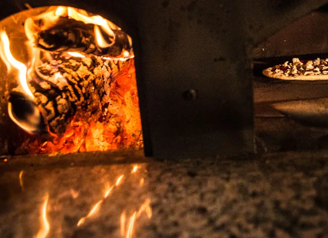 Pizza cociéndose junto a la leña encendida dentro del horno de Porco Dio